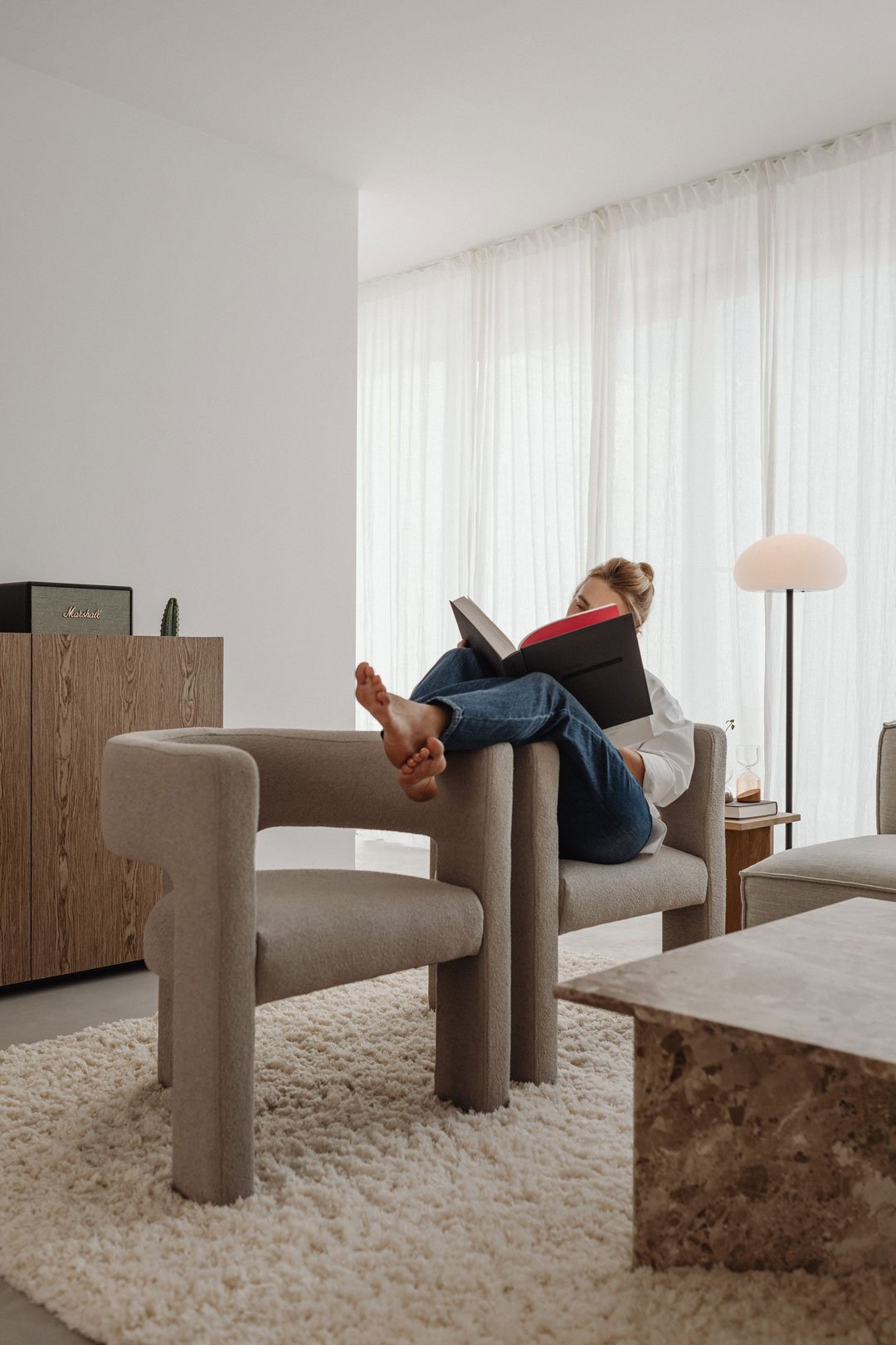 Persona leyendo en un sillón beige junto a una gran cortina blanca y luz natural.