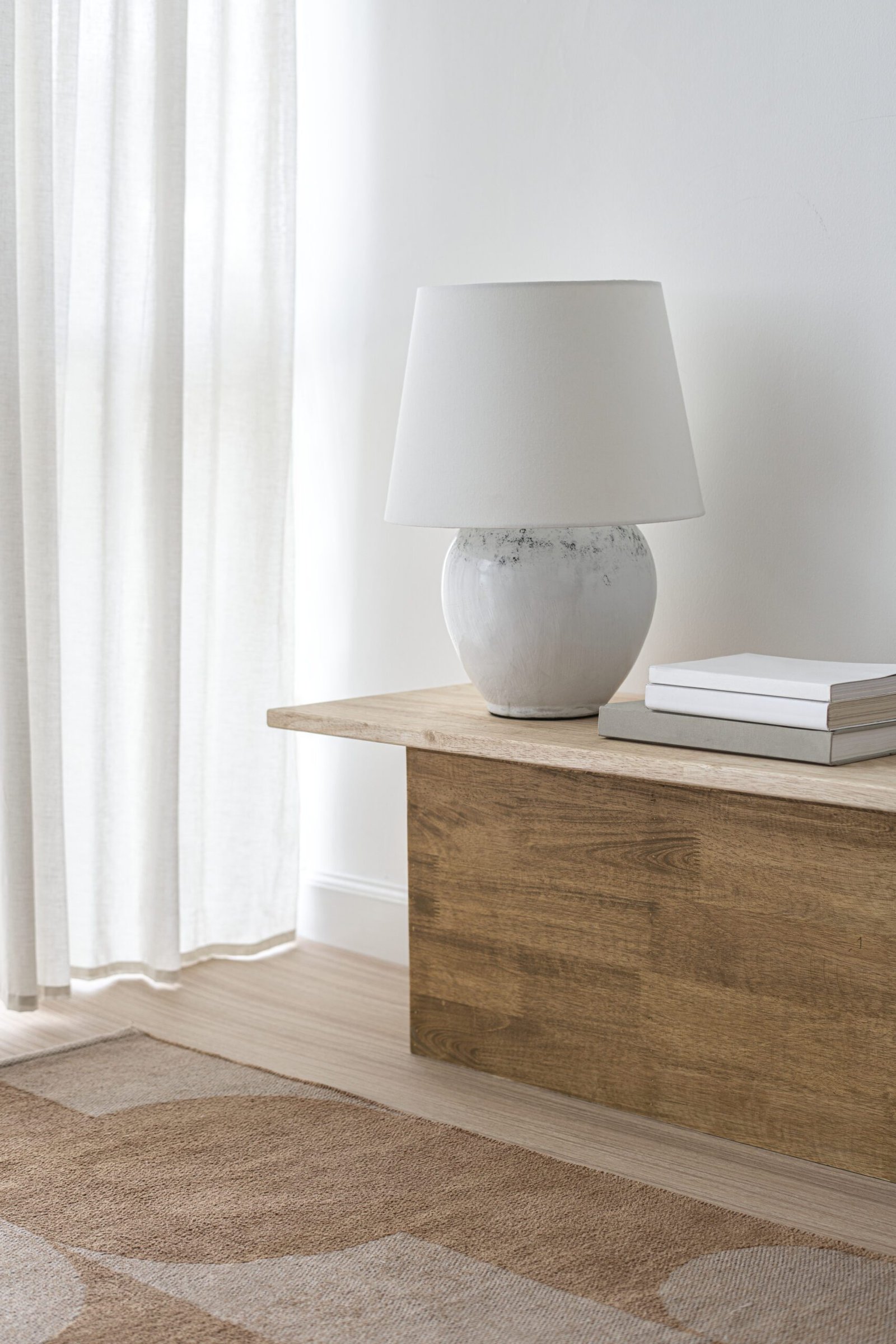 Taula de fusta amb làmpada de llum càlida al costat d’una cortina clara que filtra la llum natural